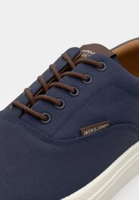 Navy canvas sneaker with brown laces and eyelets, white rubber sole, and a branded brown tag on the side. Smooth texture, casual design.