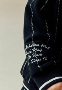 Donker gebreide trui met witte verticale strepen, met geborduurde tekst op de mouw: "Athletics Club", "Laten we Spelen" en "Sinds 82".