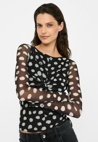 Top nero a maniche lunghe con pois bianchi, caratterizzato da un design frontale annodato e maniche trasparenti. Il materiale sembra leggero e testurizzato.