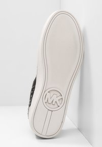 Černá podrážka tenisky s texturovaným povrchem, obsahující kruhové logo MK a bílé akcenty podél okraje, navržená pro grip a styl.