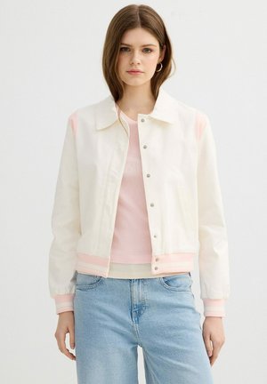 Joven mujer con chaqueta blanca con botones y detalles rosas sobre una camisa rosa y vaqueros azul claro, posando frente a un fondo blanco liso.