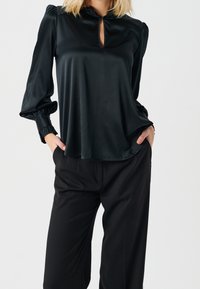 Blusa nera in satin con maniche a palloncino, dettaglio a chiave sul collo e vestibilità ampia. Abbinata a pantaloni neri su misura.