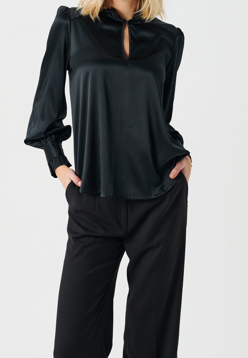 Blusa nera in satin con maniche a palloncino, dettaglio a chiave sul collo e vestibilità ampia. Abbinata a pantaloni neri su misura.