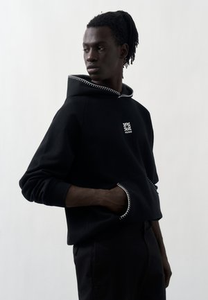 Mężczyzna z dredami w czarnej bluzie z białymi przeszyciami i logo na przodzie, patrzący na bok z jedną ręką w kieszeni.