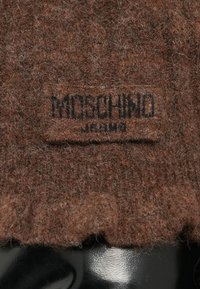 Barna gyapjú kötött anyag, texturált felülettel, amelyen egy fekete Moschino címke található a tetején. A ruhadarab alján bordás szegély található.