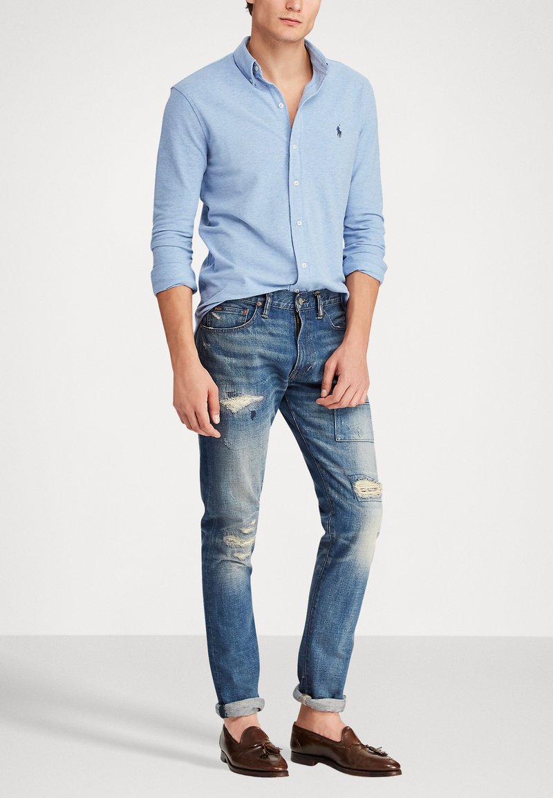 Lichtblauw overhemd met knopen, versleten blauwe jeans, bruine loafers. Het overhemd heeft een logo, de jeans hebben vervaagde plekken en opgerolde mouwen.