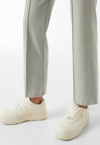 Białe sneakersy na platformie z gładkiej skóry, z perforowanym czubkiem, systemem sznurowania i minimalnym logo, zestawione z jasnoszarymi spodniami.