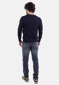 Maglione navy a maniche lunghe, orlo a coste, abbinato a jeans blu e sneakers marroni, visto da dietro su uno sfondo neutro.