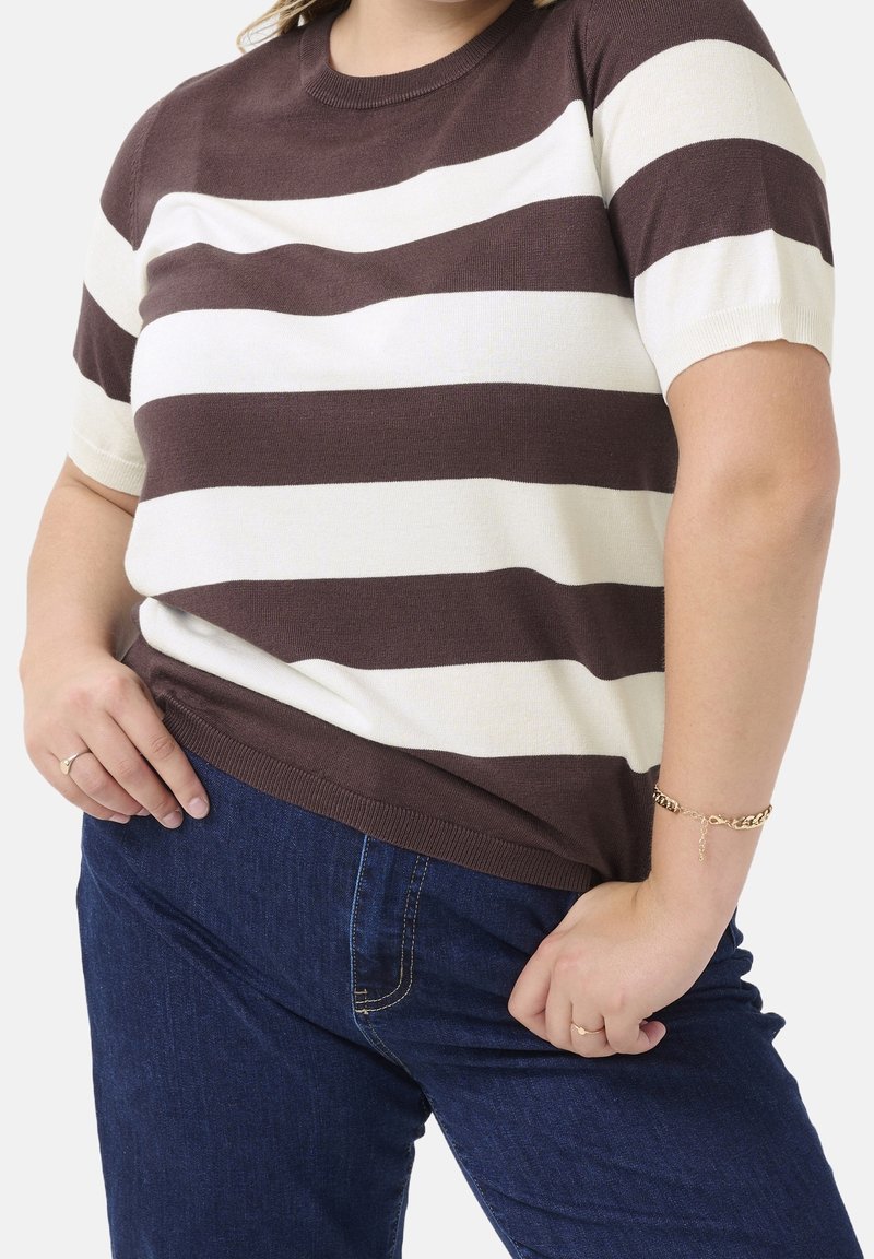 Femme portant un t-shirt à manches courtes rayé marron et blanc et un jean bleu foncé, avec un bracelet en or et des bagues, mains posées sur les hanches.