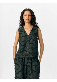 Haut sans manches vert foncé avec un décolleté en V, présentant des motifs circulaires texturés et des bords effilochés. Fermeture à cinq boutons à l'avant.