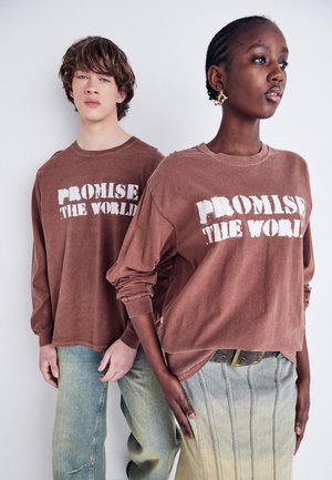 Chemises à manches longues marron avec le texte "PROMISE THE WORLD", associées à un jean bleu clair et une jupe de teinte verdâtre avec des coutures verticales.