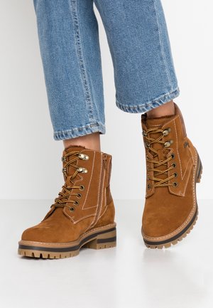 Brązowe zamszowe botki za kostkę z okrągłym noskiem, łączonym sznurowaniem i zamkiem błyskawicznym, solidną podeszwą oraz akcentami w postaci przeszyć. Zestawione z krótkimi niebieskimi dżinsami.