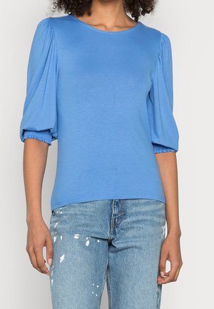Top blu corto a maniche corte con maniche a sbuffo arricciate, realizzato in tessuto morbido. Indossato con jeans azzurri chiari strappati con macchie di vernice.