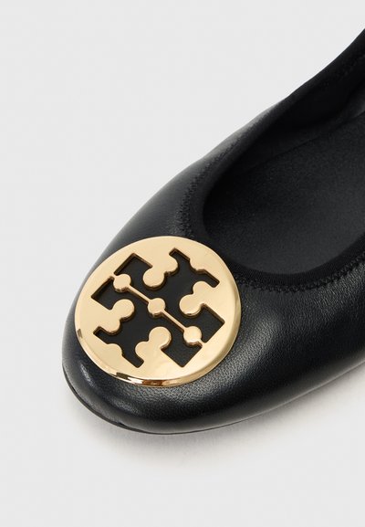 Chaussure plate en cuir noir avec un accent en logo doré rond sur le bout, dotée d'une finition lisse et d'une forme structurée et arrondie.