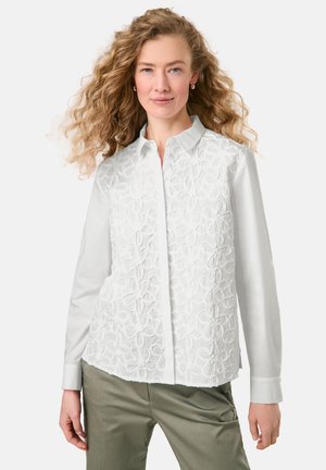 Weiße Bluse mit strukturiertem floralem Stickmuster auf der Vorderseite, langen Ärmeln, knöpfbarem Kragen und glattem Stoff. Kombiniert mit grünen Hosen.