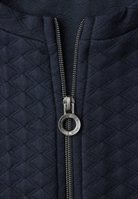 Marineblåt quiltet stof med diamantmønster, med en metallisk lynlås og en cirkulær trækflig mærket "StreetOne".