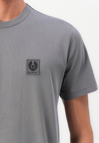 Close-up of a person wearing a grey Belstaff t-shirt with a small black logo patch on the left chest.