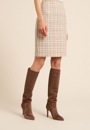 Femme portant une jupe beige à carreaux jusqu'aux genoux et des bottes marron en daim à talons hauts jusqu'aux genoux, debout sur un fond neutre.