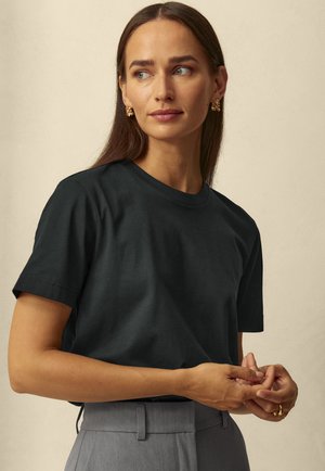 Černé bavlněné tričko s klasickým kulatým výstřihem a krátkým rukávem. Modelka má na sobě šedé kalhoty a zlaté náušnice. Měkká textura.