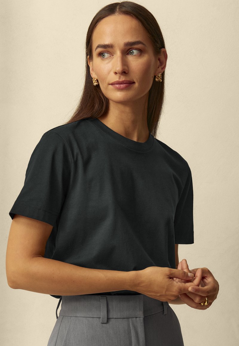 Black cotton t-shirt with a classic crew neck and short sleeves. The model wears gray trousers and gold earrings. Soft texture.
