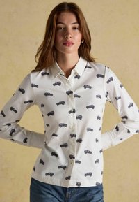 Biela košeľa na gombíky s opakujúcim sa čiernym vzorom vintage áut, dlhé rukávy a klasický golier. Stylizovaná s modrými džínsy.