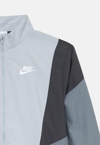 Nike jakna s vjetrom koja ima zatvaranje na zip, visoki ovratnik, blokiranje boja u sivoj i crnoj, glatku teksturu i istaknutu bijelu oznaku.