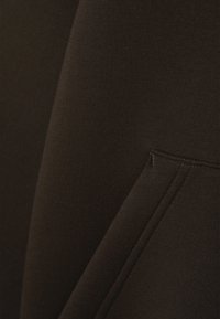 Tecido castanho-escuro com uma textura suave. Apresenta um bolso lateral com costura visível e um leve brilho, sugerindo um design confortável.