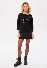 Zwarte top met lange mouwen en een vlinder van pailletten, gecombineerd met een zwarte denim rok en veterlaarzen. Soepele textuur, losse pasvorm.