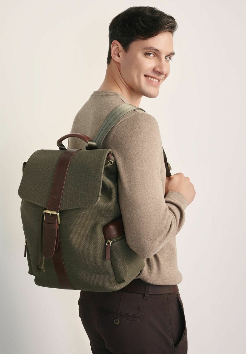 Młody mężczyzna w beżowym swetrze i ciemnych spodniach uśmiecha się, nosząc zielony plecak z brązowym skórzanym paskiem na jednym ramieniu.