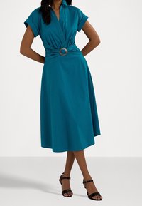 Teal bomullsklänning med krage, vikdesign och en dekorativ bältesspänne. Utställd kjol som når mitt på vaden, kombinerad med svarta klackar.