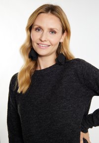 Fekete kötött pulóver kerek nyakkivágással, texturált anyagból, hosszú ujjakkal, nagy fekete virág alakú fülbevalóval viselve.