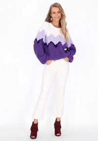 Pulover colorat din tricot cu un model în zigzag în nuanțe de purpuriu și alb, având accente decorative negre și purpurii. Asortat cu blugi albi evazați.