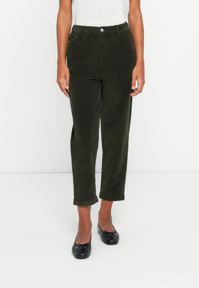 Femme portant un pantalon en velours côtelé vert foncé avec les revers roulés et des ballerines noires, debout devant un fond blanc.