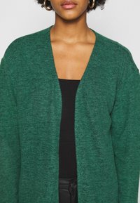 Zielony sweter z dzianiny, z głębokim dekoltem w kształcie litery V i otwartym przodem, zaprojektowany w miękkiej teksturze. Noszony na czarnym dopasowanym topie.