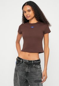 Καφέ cropped μπλούζα με κοντά μανίκια και ριμπ υφή, με μπλε λογότυπο "HUGO". Συνδυασμένη με γκρι τζιν με φθορές και μαύρη ζώνη.