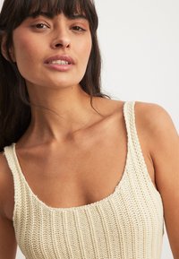 Neuleliivi off-white värviga, millel on tekstureeritud ribakujundus, laiad rihmad ja ruudukujuline kaelus. Pehme ja kerge materjal mugavuseks.