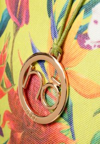 Gouden ronde hanger met het "nö" logo, bevestigd op een stoffen achtergrond met een bloemendessin in levendige kleuren.
