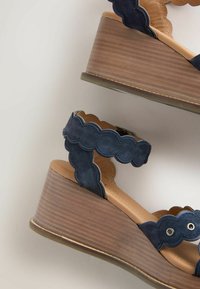 Sandales compensées en daim bleu marine à bord festonné, talons plateforme en bois et brides de cheville avec œillets métalliques sur un fond clair.
