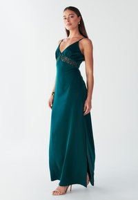 Abito lungo in satin color turchese con profondo scollo a V. Presenta dettagli in pizzo in vita e uno spacco alto sulla coscia su un lato. Spalline sottili, texture liscia.