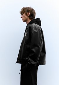 Zwarte leren jas met een capuchon, voorzien van een ritssluiting, twee zijzakken en een opvallend logo op de borst. Soepele textuur.