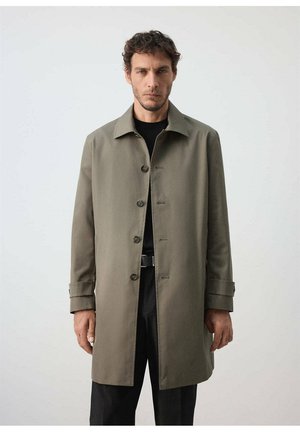 Hombre con un abrigo verde oliva con botones hasta la rodilla sobre una camisa negra y pantalones negros, con un fondo liso y claro.