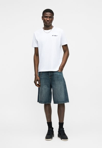 Άνδρας στέκεται φορώντας λευκό κοντομάνικο πουκάμισο με μικρό λογότυπο, τζιν σορτς μέχρι το γόνατο, μαύρες κάλτσες και μαύρα αθλητικά παπούτσια, σε λευκό φόντο.