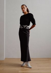 Black top with puff sleeves, fitted black skirt, and a patterned white belt. Shoes in a snake print design. Light wooden floor background.