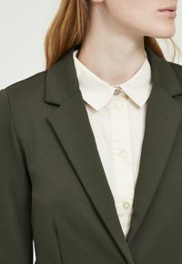 Olive green blazer with a structured lapel and cream button-up shirt underneath. Features a decorative button at the collar and smooth texture.