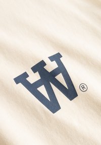 T-shirt beige clair présentant un grand logo bleu foncé avec des lettres stylisées "W" et le symbole de marque déposée. Tissu doux et texturé.