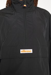 Czarna kurtka przeciwdeszczowa z wysokim kołnierzem, pół-zamkiem z przodu oraz pomarańczowym, haftowanym logo "ellesse" na białej łatce. Lekka, gładka tekstura.