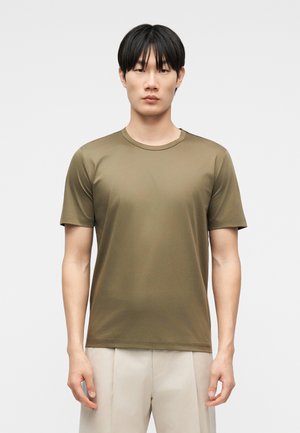Un homme portant un t-shirt à manches courtes olive à col rond et un pantalon beige, debout devant un fond gris clair uni.