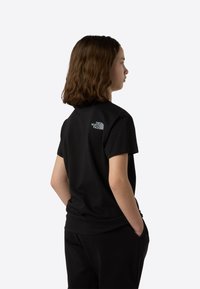 Črna bombažna majica s kratkimi rokavi in okrolim izrezom, na zgornjem delu hrbta majhen siv logotip. Nosijo jo s črnimi hlačami, model ima hrbet obrnjen stran.