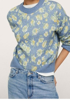 Personne portant un pull en maille bleu à motif floral sur une chemise blanche, associé à un jean denim bleu clair, les mains dans les poches arrière.