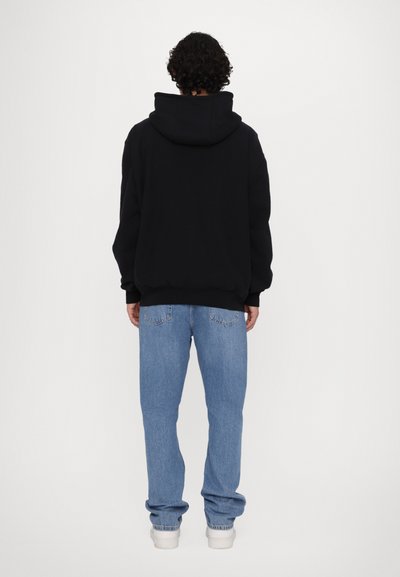 Hoodie noir avec une capuche spacieuse, des poignets côtelés, associé à un jean en denim bleu clair ample et des baskets blanches. Design minimaliste.
